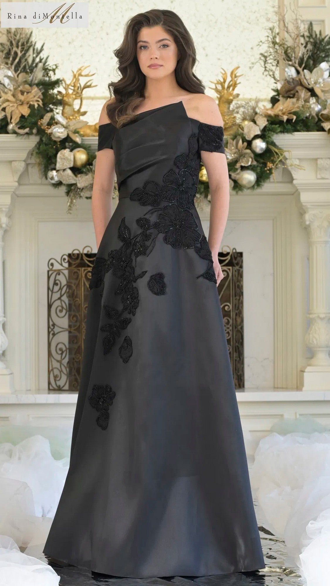A woman stands indoors wearing a black Rina Di Montella RD3344 long formal dress with off-the-shoulder and floor-length floral appliqué details. White and gold holiday decorations are visible in the background.